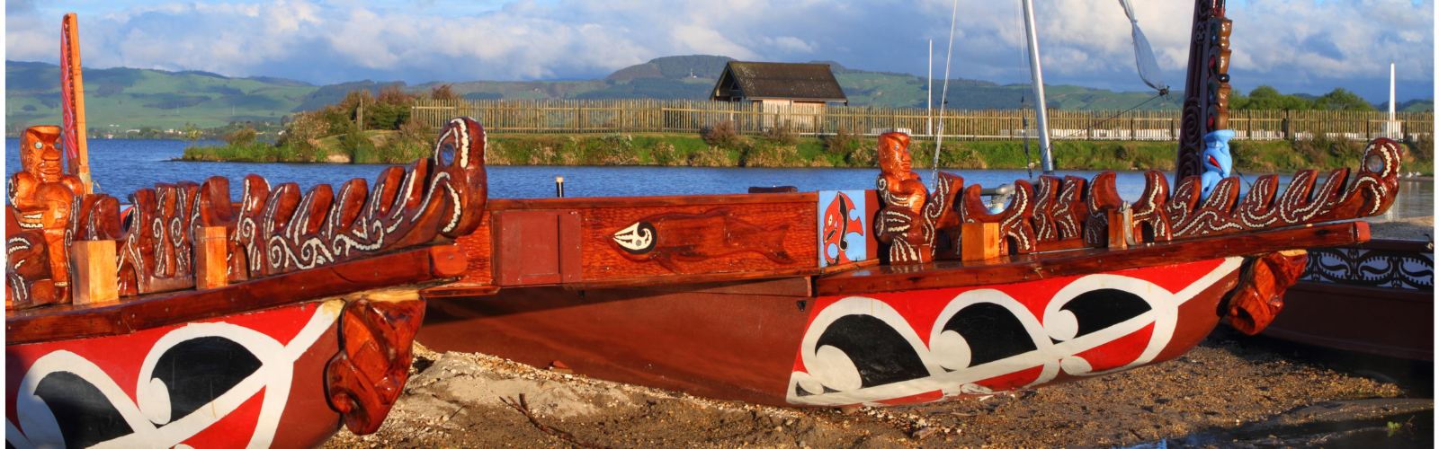 Two wooden boats painted mostly red with other designs in white sit on the rocky shore next to a river. 